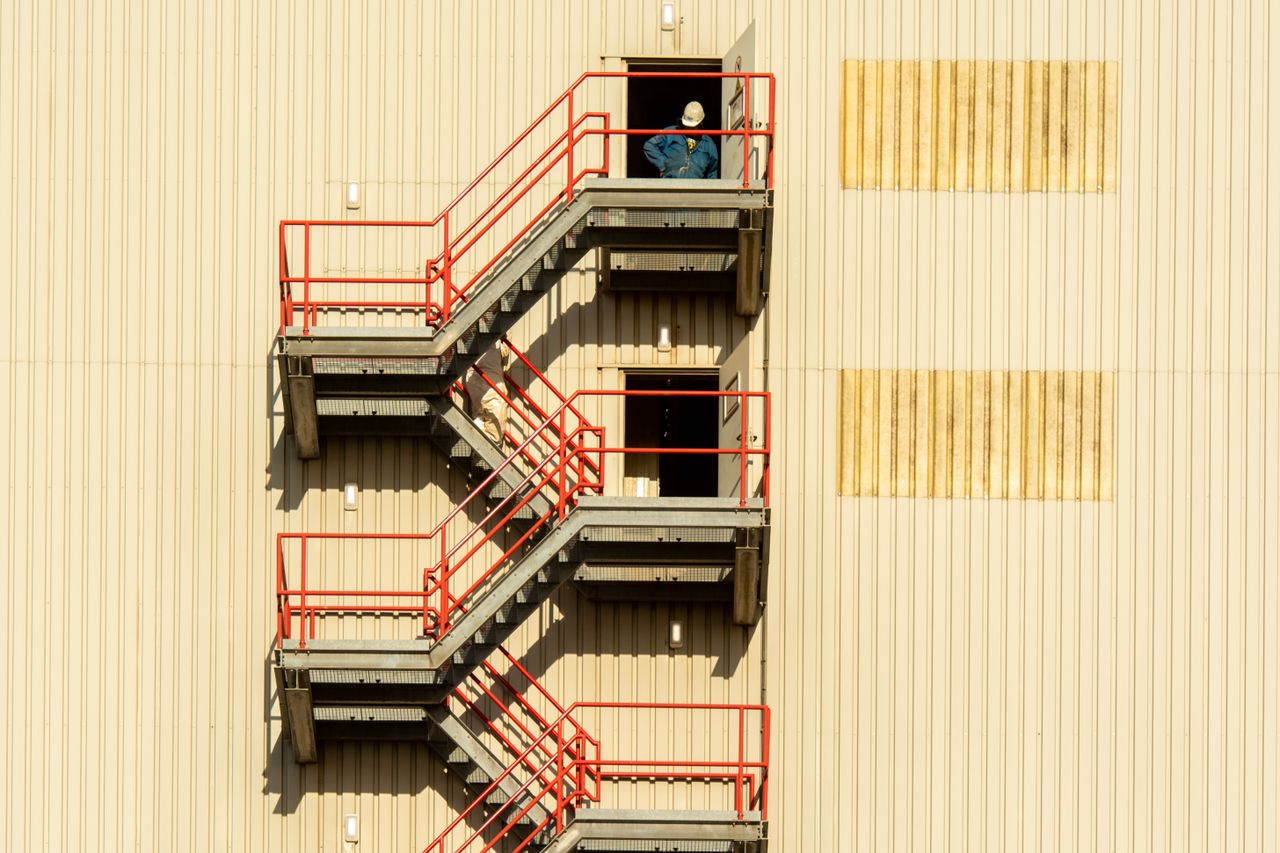 Home Worker in hardhat standing on fire escape stairs of industrial building with beige walls.
